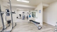 Located in Saint Louis, MO, this fitness center offers a clean, modern exercise environment designed to support a well-rounded workout routine. The space is spread across a single floor and is organized into distinct zones that accommodate both cardio and strength training activities.

The cardio area features a row of elliptical trainers and additional aerobic machines, all positioned beneath flush-mounted skylight panels that flood the room with bright, even lighting. A wall-mounted flat-screen television is conveniently positioned for use during cardio sessions. Light wood-style flooring runs throughout, contributing to the facility's fresh, contemporary aesthetic.

The strength training area is equally well-appointed, offering a multi-station cable machine, an adjustable weight bench, a dumbbell rack with a full set of free weights, and a power tower for bodyweight exercises. A large wall mirror expands the visual depth of the space and allows users to monitor their form during workouts.

Additional amenities include a stability ball, a wall-mounted paper towel dispenser, and a keypad entry system for secure, controlled access. The crisp white walls and neutral finishes create a bright, motivating atmosphere throughout. The layout flows naturally between training zones, making efficient use of the available floor area while maintaining an open, uncluttered feel.