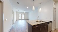 This well-appointed single-floor unit offers a thoughtfully designed open-concept layout that seamlessly connects the kitchen, dining, and living areas. The kitchen is a standout feature, showcasing dark espresso cabinetry, light granite countertops, a central island with a stainless steel undermount sink, and pendant lighting overhead. Stainless steel appliances — including a refrigerator, electric range, microwave, and a tile backsplash — complete the functional and stylish cooking space.

The living area is generously proportioned, bathed in natural light streaming through French doors that open to the outdoors. Light hardwood-style flooring flows throughout the main living areas, complemented by recessed lighting and a ceiling fan. The bedroom features plush carpeting, a ceiling fan, and large windows with blinds that allow for ample natural light while maintaining privacy.

The bathroom continues the high-end aesthetic with espresso cabinetry, granite countertops, a wide mirror, and wood-style flooring. An adjacent walk-in closet provides substantial storage space. Throughout the unit, crisp white walls, clean architectural lines, and consistent finishes create a bright, modern, and cohesive living environment.