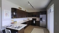 This single-floor space showcases a thoughtfully designed kitchen that balances style and functionality. The kitchen is defined by rich, dark-toned cabinetry that extends to the upper walls, providing generous storage throughout. The cabinetry is complemented by crisp white countertops, creating a striking contrast that gives the space a clean, contemporary aesthetic.

A built-in wine rack with an X-pattern design adds a distinctive decorative element near the stainless steel French door refrigerator with an in-door water and ice dispenser. The cooking area features a black electric range and an over-the-range microwave, keeping the appliance suite cohesive. An undermount sink is neatly positioned beneath a set of transom-style windows, allowing natural light to filter into the space.

Track lighting installed along the ceiling provides adjustable, focused illumination across the work surfaces. Open shelving on one wall offers both display and accessible storage. The kitchen extends into a peninsula with seating, making it a practical and social hub. Light hardwood-style flooring and a patterned area rug tie the space together with warmth.