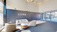The Leora building lobby at 2125 Rue Ward in Montréal offers a sophisticated and contemporary first impression. The space is defined by its dramatic double-height ceilings, which amplify the sense of openness and grandeur. Floor-to-ceiling glazing along the entrance façade floods the interior with abundant natural light and frames views of the surrounding streetscape, creating a seamless connection between indoors and outdoors.

The main lounge area features a carefully curated seating arrangement, including a plush white sofa, rounded upholstered armchairs, and a sleek wooden coffee table, all set atop a deep grey area rug that anchors the space. The signature Leora branding is prominently displayed on a bold navy-blue feature wall, lending the lobby a strong sense of identity and elegance. Light wood flooring complements the crisp white walls and clean architectural lines throughout.

A sculptural pendant light fixture adds a refined artistic touch overhead, while wall-mounted and recessed lighting provide warm, layered illumination. Transitioning deeper into the building, the hallway continues the refined palette with grey walls, large-format porcelain tile flooring, carpeted corridors, and subtle recessed ceiling lighting, guiding residents toward the elevators and residential wings with understated sophistication.