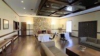 Located at 4950 Willow Creek Dr in Jackson, Mississippi, this single-floor gathering space combines classic architectural charm with warm, inviting design elements. The centerpiece of the main hall is a dramatic floor-to-ceiling stone fireplace, providing a rustic yet elegant focal point that anchors the room. Overhead, a distinctive coffered ceiling with mirrored inlays adds depth and a touch of sophistication, reflecting light throughout the space and enhancing its sense of openness. Rich hardwood flooring runs throughout, complementing the warm, neutral wall tones accented by dark wood trim. Recessed lighting and tall floor lamps provide layered illumination, creating a welcoming ambiance suitable for a variety of events. The layout comfortably accommodates round tables dressed in white linens alongside lounge seating, making it well-suited for receptions, gatherings, or private functions. Natural light filters in through windows, contributing to the bright and airy atmosphere. The combination of the stone fireplace, mirrored coffered ceiling, and classic interior finishes gives this space a timeless character, offering a versatile and memorable setting for events of all kinds.