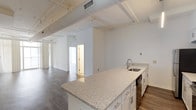 This single-floor unit offers a clean, contemporary design characterized by an open layout, bright white walls, and rich hardwood-style flooring throughout the main living areas. The open-concept kitchen serves as a central feature, showcasing a large granite-topped island with an undermount sink, white shaker-style cabinetry, a subway tile backsplash, and a full suite of stainless steel appliances including a refrigerator, electric range, and microwave. A modern linear pendant light illuminates the kitchen space. Exposed cylindrical ductwork and structural beams run across the ceilings, lending an industrial character to the otherwise clean aesthetic. The living and bedroom areas are generously proportioned, with floor-to-ceiling windows and vertical blinds that invite abundant natural light and offer views of the surrounding urban environment. The bedrooms feature hardwood-style flooring and dedicated closet space. The bathrooms are finished with granite-topped white vanities, framed mirrors, and classic mosaic tile flooring. One bathroom includes a full tub-shower combination, while both are appointed with brushed nickel fixtures and hardware. Overall, the space presents a well-balanced blend of industrial and modern design elements.