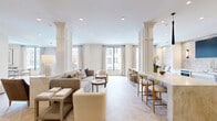 Located at 3551–3571 Scotts Ln in Philadelphia's 19129 neighborhood, this thoughtfully designed single-floor amenity space offers a generous, open layout ideal for gathering and socializing. The interior is defined by a sophisticated neutral palette of creams, whites, and warm taupes that lend a calm, refined atmosphere throughout.

Striking herringbone-patterned hardwood flooring flows across the main areas, complemented by recessed lighting and clean white ceilings. Structural columns with paneled detailing add architectural character while subtly defining distinct zones within the open floor plan. Multiple seating arrangements are distributed throughout the space, including plush sofas, accent chairs, and built-in banquette seating, providing versatile areas for relaxation and conversation.

A well-appointed kitchenette runs along one wall, featuring white shaker cabinetry, a light stone countertop, an undermount sink, stainless steel appliances including a built-in microwave and dishwasher, and a textured stone tile backsplash. A wall-mounted flat-screen television and flanking sconce lighting anchor this functional area. A bar-height counter with barstool seating offers an additional casual gathering spot.

Large windows throughout invite abundant natural light and provide views of the surrounding neighborhood. Curated artwork adorns the walls, enhancing the polished, welcoming character of this well-appointed communal space.
