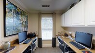 This single-floor space functions as a dedicated business center designed to support productivity in a comfortable, professional environment. The room features two built-in workstations positioned along opposing walls, each equipped with a full desktop computer setup, keyboard, and ergonomic office chair, providing ample workspace for multiple users simultaneously. Above one of the desks, white upper cabinetry offers generous storage, keeping the area organized and clutter-free. A centrally placed window dressed with elegant two-toned drape panels and white horizontal blinds allows natural light to filter into the room, creating a bright and welcoming atmosphere while maintaining privacy. The warm beige walls are accented by crisp crown molding, lending the space a polished, refined character. A framed abstract canvas adds a touch of color and artistic flair to the room. The neutral-toned carpet complements the overall aesthetic, contributing to a calm and focused work environment. The thoughtful layout maximizes functionality within the space, making it a practical and inviting setting for business tasks, remote work, or general computing needs.