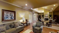 This single-floor space offers a welcoming and well-appointed interior defined by its open, flowing layout and warm neutral color palette. The living area serves as the central hub, featuring hardwood flooring that extends throughout and crown molding that adds a refined architectural touch. Recessed lighting is installed throughout the ceiling, complementing the warm ambient glow created by accent lamps and built-in lighting details.

The main seating area is generously sized, accommodating a plush sofa and coordinating upholstered armchair arranged around a patterned area rug. A wooden side table with a decorative lamp adds character to the space. Decorative columns frame the transition into an adjacent area, which includes a fireplace with a classic white mantle, creating a focal point that enhances the overall warmth of the room.

Further into the space, a secondary seating or work area is visible, along with a glass-top table and additional furnishings. Multiple doorways lead to other areas of the unit, suggesting a practical and well-connected floor plan. The overall ambiance is cozy yet sophisticated, blending traditional design elements with comfortable, livable furnishings.