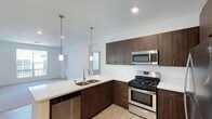This single-floor space showcases a thoughtfully designed open-concept layout, with the kitchen serving as a central highlight. The kitchen features rich, dark-toned flat-front cabinetry that provides ample storage, complemented by crisp white countertops that create a striking contrast. Stainless steel appliances, including a gas range, built-in microwave, and dishwasher, lend a polished, contemporary feel throughout.

A double-basin sink is positioned along the kitchen peninsula, which opens directly toward the adjoining living area, promoting a connected and social atmosphere. Two pendant lights hang above the peninsula, adding focused task lighting while contributing to the modern aesthetic.

The space benefits from recessed ceiling lighting and an abundance of natural light streaming in through windows visible in the background, keeping the interior bright and airy. Light-toned flooring runs throughout, further enhancing the sense of openness. The clean lines, neutral palette, and seamless transition between the kitchen and living area give this single-floor space a cohesive, contemporary character well-suited for everyday living and entertaining.