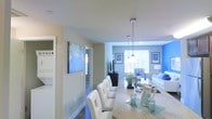This single-floor residence features an open-concept layout that connects the dining and living areas in one cohesive, light-filled space. The dining area is anchored by a granite-style countertop bar with tufted white chairs and illuminated by sleek pendant lights with chrome finishes that hang from smooth white ceilings. A large abstract blue canvas artwork adds a bold visual statement near the kitchen corridor, complementing the overall cool-toned color palette throughout.

The living area beyond is bright and welcoming, featuring large windows that allow ample natural light to fill the room. Blue accent walls and blue-and-white soft furnishings create a cohesive coastal-inspired aesthetic. A white sofa, glass-top coffee table, and decorative greenery contribute to the clean, modern feel of the space.

A hallway visible from the dining area leads to additional rooms, hinting at further functional space beyond what is immediately visible. The dark hardwood-style flooring grounds the interior while contrasting elegantly with the light walls and furnishings, giving the overall space a polished, contemporary character.