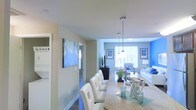 This single-floor residence features an open-concept layout that connects the dining and living areas in one cohesive, light-filled space. The dining area is anchored by a granite-style countertop bar with tufted white chairs and illuminated by sleek pendant lights with chrome finishes that hang from smooth white ceilings. A large abstract blue canvas artwork adds a bold visual statement near the kitchen corridor, complementing the overall cool-toned color palette throughout.

The living area beyond is bright and welcoming, featuring large windows that allow ample natural light to fill the room. Blue accent walls and blue-and-white soft furnishings create a cohesive coastal-inspired aesthetic. A white sofa, glass-top coffee table, and decorative greenery contribute to the clean, modern feel of the space.

A hallway visible from the dining area leads to additional rooms, hinting at further functional space beyond what is immediately visible. The dark hardwood-style flooring grounds the interior while contrasting elegantly with the light walls and furnishings, giving the overall space a polished, contemporary character.