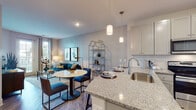 This single-floor residence is designed with a contemporary open-concept layout that seamlessly connects the kitchen, dining, and living areas. The kitchen is well-appointed with shaker-style cabinetry in a soft gray finish, granite countertops, a stainless steel undermount sink, and a full suite of stainless steel appliances including a range, over-the-range microwave, and refrigerator. A granite-topped island with pendant lighting overhead provides additional prep space and a casual dining option.

The open living and dining area features wood-style plank flooring throughout and flows naturally toward large windows and a glass-paned exterior door, allowing generous natural light and outdoor views. The living room accommodates a comfortable seating arrangement, complemented by neutral wall tones and tasteful accent lighting.

The bedrooms offer plush carpeting, ceiling fans, and soft teal accent walls that create a calm, restful atmosphere. Windows dressed with patterned curtains provide privacy while still welcoming natural light. The bathroom features gray shaker-style cabinetry, a granite-topped vanity with a framed mirror, wood-style flooring, and clean, bright fixtures—delivering a cohesive and polished finish consistent throughout the residence.