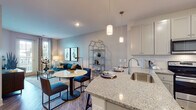 This single-floor residence is designed with a contemporary open-concept layout that seamlessly connects the kitchen, dining, and living areas. The kitchen is well-appointed with shaker-style cabinetry in a soft gray finish, granite countertops, a stainless steel undermount sink, and a full suite of stainless steel appliances including a range, over-the-range microwave, and refrigerator. A granite-topped island with pendant lighting overhead provides additional prep space and a casual dining option.

The open living and dining area features wood-style plank flooring throughout and flows naturally toward large windows and a glass-paned exterior door, allowing generous natural light and outdoor views. The living room accommodates a comfortable seating arrangement, complemented by neutral wall tones and tasteful accent lighting.

The bedrooms offer plush carpeting, ceiling fans, and soft teal accent walls that create a calm, restful atmosphere. Windows dressed with patterned curtains provide privacy while still welcoming natural light. The bathroom features gray shaker-style cabinetry, a granite-topped vanity with a framed mirror, wood-style flooring, and clean, bright fixtures—delivering a cohesive and polished finish consistent throughout the residence.