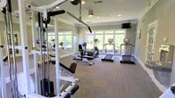This fitness center is thoughtfully designed to provide a comprehensive workout environment in a bright, welcoming setting. The space is outfitted with a range of cardio equipment, including multiple treadmills and an elliptical machine, as well as a stationary bike and cable-based strength training apparatus, offering a well-rounded selection for users of all fitness levels. A wall of floor-to-ceiling mirrors lines one side of the room, visually expanding the space and allowing for proper form checks during exercise. The flooring consists of durable, textured carpet tiles that provide comfort underfoot while anchoring the equipment securely. Large, multi-pane windows span the far wall, flooding the interior with natural light and offering pleasant views of the surrounding greenery, creating an inviting and energizing atmosphere. A wall-mounted flat-screen television adds an entertainment option for those looking to stay engaged during longer sessions. Recessed lighting in the white ceiling ensures the space remains well-lit at all times. The neutral gray walls contribute to a clean, modern aesthetic, while a ceiling fan supports air circulation throughout. Overall, this fitness center balances functionality with a comfortable, airy ambiance.