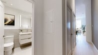 This thoughtfully designed single-floor unit maximizes its compact footprint with an open, modern layout and high-quality finishes throughout. The combined kitchen and dining area serves as the heart of the space, featuring a well-appointed kitchen with grey shaker-style cabinetry, glass-front upper cabinets, a marble-look tile backsplash, stainless steel appliances including a range and built-in microwave, and under-cabinet lighting. A dark wood dining table anchors the space and flows naturally into a living area outfitted with light hardwood flooring and soft, neutral-toned walls. A dedicated workspace with a window offering urban skyline views adds functional versatility to the living area. The hallway connects the main living space to the bathroom and features a clean, white palette with light wood flooring continuing throughout. The bathroom is a standout, showcasing large-format grey stone-look tiles, a floating dual vanity with a white countertop and backlit mirror, and a frameless glass-enclosed walk-in shower with built-in niches. The overall aesthetic is cohesive, contemporary, and refined — emphasizing clean lines, neutral tones, and purposeful design at every turn.