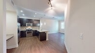 This contemporary 610 square foot apartment, located in The Residences At Bella Terra in Huntington Beach, California, offers a thoughtfully designed living space. The single-floor layout encompasses one bedroom, one bathroom, and a range of functional areas.

The kitchen stands out with its modern aesthetic, featuring dark wood cabinetry contrasting beautifully with stainless steel appliances. A mosaic tile backsplash adds visual interest, while the light-colored quartz countertops provide ample workspace. The kitchen's L-shape design maximizes efficiency and includes a built-in desk area, perfect for a small home office setup.

The bedroom, measuring 130 square feet, offers a cozy retreat with plush carpeting and a large window that allows natural light to flood the space. A ceiling fan ensures comfort year-round. The bathroom continues the modern theme with a spacious vanity, large mirror, and a shower/tub combination.

One of the apartment's highlights is the in-unit laundry, discreetly tucked away behind closet doors, offering the convenience of at-home laundry facilities. The hallway provides access to additional storage spaces.

Throughout the apartment, you'll find a cohesive design with neutral wall colors, recessed lighting, and a mix of carpeting in the bedroom and living areas, with wood-look flooring in the kitchen and bathroom. This thoughtful blend of materials creates a harmonious and inviting living environment, perfect for single professionals or couples seeking a contemporary urban lifestyle.