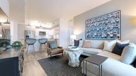 This single-floor residence showcases a cohesive contemporary design with a carefully curated color palette of soft blues, warm grays, and crisp whites carried throughout. The open-concept living area features light wood-style flooring, a large sliding glass door that invites natural light and provides access to an outdoor balcony, and a neutral sectional sofa anchored by a striking organic wood coffee table. A large blue botanical artwork serves as a focal point on the accent wall, complementing the space's coastal-modern aesthetic.

The kitchen is outfitted with high-gloss white cabinetry, a white subway tile backsplash, stainless steel appliances, and a generous island with quartz countertops and pendant lighting overhead — seamlessly connecting to the living area for an open, functional layout.

The bedroom features a bold deep-teal accent wall, a ceiling fan, light wood flooring, and ample natural light through a window dressed with sheer white curtains. A king-sized upholstered bed with layered neutral bedding anchors the room.

Both bathrooms are finished with light wood-tone floating vanities, white quartz countertops, large mirrors, and wall sconces, with light wood-style flooring maintaining design consistency throughout the residence.