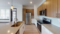 This contemporary single-floor residence showcases a thoughtfully designed layout with clean lines, crisp white walls, and light wood-tone flooring that flows seamlessly throughout the space. Dark-trimmed doorframes and bold interior doors provide a striking contrast against the bright, neutral palette, lending the space a modern, polished character.

The kitchen is a standout feature, equipped with a full suite of stainless steel appliances including a refrigerator, electric range, microwave, and dishwasher. A two-tone cabinetry scheme pairs warm wood upper cabinets with white lower cabinets, complemented by a classic subway tile backsplash and stone-finish countertops. A deep undermount sink with a modern faucet anchors the kitchen island, which provides additional prep and dining space.

The bedroom is bright and generously proportioned, with a large window that invites natural light and offers views of the surrounding area. The open layout provides flexibility for a variety of furniture arrangements.

The bathroom features a white vanity with stone countertops, a tub-shower combination, and consistent use of modern hardware and fixtures. An adjoining hallway leads between the bathroom and bedroom, offering convenient flow.

Recessed lighting throughout ensures well-lit interiors, while the cohesive design choices create an inviting, contemporary atmosphere ideal for comfortable everyday living.
