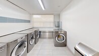 Located at 66 Spadina Rd in Toronto's Annex neighbourhood, this well-appointed shared laundry facility offers a clean, functional, and comfortable space for residents. The room is finished with bright white walls and large-format light-coloured floor tiles, giving the space a clean and modern aesthetic throughout.

The laundry area is equipped with a row of front-loading, commercial-grade stainless steel washing machines and dryers lined along the walls, accented by light blue tile detailing that adds a touch of colour to the otherwise crisp white interior. Stainless steel countertops provide ample surface area for folding and sorting laundry, while overhead fluorescent and recessed lighting ensures the space is well-lit at all times.

A dedicated waiting lounge area offers residents a comfortable place to relax, featuring a wall-mounted flat-screen television, a cushioned armchair, and a wooden coffee table — making the time spent waiting for laundry cycles more enjoyable. A window allows natural light to filter into this seating area.

Additionally, the facility includes a Canada Post parcel locker station, providing residents with a convenient and secure package pickup solution. A community bulletin board and folding table complete the practical amenities of this thoughtfully designed shared space.