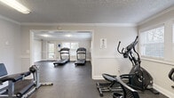 This fitness center is organized across a single floor and divided into two interconnected rooms, offering a functional and comfortable workout environment. The space features durable dark rubber flooring throughout, textured ceilings with crown molding, and neutral-toned walls accented by a classic chair rail detail — lending a clean, polished aesthetic to the facility.

The cardio zone is home to multiple treadmills positioned near large windows that invite natural light and provide outdoor views, creating an energizing atmosphere for exercise. An elliptical trainer and a rowing machine round out the cardio equipment selection, catering to a variety of low-impact workout preferences.

The adjoining strength training area is equipped with a multi-station cable machine, a weight bench, a stationary bike, and additional cross-training equipment, providing users with comprehensive options for resistance and circuit training. Interior doorways connect the two zones seamlessly, allowing for easy movement throughout the facility.

Each room benefits from flush-mounted ceiling lighting to supplement natural light. Entry points with glass-paneled doors open to an outdoor area, enhancing ventilation and accessibility. Informational signage is posted throughout the space. Overall, the fitness center presents a well-maintained, multi-purpose exercise facility suited for a broad range of fitness routines.