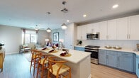 This single-floor residence is designed with a contemporary open-concept layout that seamlessly connects the kitchen, dining, and living areas. The kitchen is a standout feature, showcasing a two-tone cabinet design pairing crisp white upper cabinets with soft blue-grey lower cabinets, all complemented by white quartz countertops. A generously sized island with seating for multiple guests serves as a natural gathering point, illuminated by globe pendant lights overhead. Stainless steel appliances, including a gas range, microwave, and refrigerator, complete the functional kitchen setup. Light wood-look flooring flows throughout the main living areas, adding warmth and continuity.

The living room offers a relaxed, open atmosphere, featuring a ceiling fan, neutral warm-toned walls, and windows that bring in natural light. The bedroom is comfortably sized with plush carpet, a ceiling fan, and a window with outdoor views.

The bathroom is notably well-equipped, featuring a dual-sink vanity with the same blue-grey cabinetry found in the kitchen, a white quartz countertop, backlit mirrors, and a full tub-shower combination with tile surround. The cohesive design choices throughout create a polished, modern ambiance.