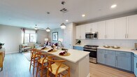 This single-floor residence is designed with a contemporary open-concept layout that seamlessly connects the kitchen, dining, and living areas. The kitchen is a standout feature, showcasing a two-tone cabinet design pairing crisp white upper cabinets with soft blue-grey lower cabinets, all complemented by white quartz countertops. A generously sized island with seating for multiple guests serves as a natural gathering point, illuminated by globe pendant lights overhead. Stainless steel appliances, including a gas range, microwave, and refrigerator, complete the functional kitchen setup. Light wood-look flooring flows throughout the main living areas, adding warmth and continuity.

The living room offers a relaxed, open atmosphere, featuring a ceiling fan, neutral warm-toned walls, and windows that bring in natural light. The bedroom is comfortably sized with plush carpet, a ceiling fan, and a window with outdoor views.

The bathroom is notably well-equipped, featuring a dual-sink vanity with the same blue-grey cabinetry found in the kitchen, a white quartz countertop, backlit mirrors, and a full tub-shower combination with tile surround. The cohesive design choices throughout create a polished, modern ambiance.