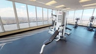 This fitness center is defined by its expansive floor-to-ceiling window walls that wrap around multiple sides of the space, flooding the interior with natural light and offering striking elevated panoramic views of the surrounding cityscape, river, and treetops beyond. The open layout is organized across a single level, with durable rubber flooring throughout that provides cushioning and traction suitable for a range of exercise activities.

The cardio zone features treadmills by True Fitness, an elliptical trainer, and a stationary bike, all positioned to take full advantage of the remarkable outdoor views. A wall-mounted television screen is available for entertainment during cardio sessions. The strength training area includes a Life Fitness multi-station cable machine paired with an adjustable weight bench, offering a versatile range of resistance exercises. A dumbbell rack with multiple weight increments and a yoga mat provide additional free-weight and flexibility training options.

The space also includes a Pepsi vending machine for post-workout refreshment and a fire safety station along one wall. A tiled entryway visible through sliding glass doors connects the fitness area to adjacent building corridors. The drop ceiling with recessed fluorescent panel lighting complements the abundant natural light. Overall, the fitness center presents a bright, functional, and well-appointed environment ideal for a comprehensive workout routine.