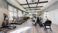 Located at Windsor Cay Blvd in Clermont, Florida, this fitness center offers a generously sized, single-floor exercise room designed to accommodate a wide range of workout routines. The space is architecturally distinguished by a coffered ceiling with dark wood beams and large industrial ceiling fans, lending the room a polished yet functional aesthetic. Recessed lighting complements the abundant natural light streaming in through multiple large windows that line the walls, creating a bright and energizing atmosphere.

The flooring throughout is a durable, wood-look surface in neutral tones, adding warmth to the space while remaining practical for high-traffic fitness use. The exercise room is divided into clearly defined zones: a cardio section houses a row of treadmills and elliptical machines, while a dedicated strength area features a power rack, adjustable benches, a full dumbbell rack, and multiple cable and weight machines. Cycling bikes are also available for low-impact cardio options.

Floor-to-ceiling mirrors on one wall enhance the sense of depth and allow users to monitor their form during strength training. Wall-mounted televisions provide entertainment throughout the workout. A separate, more intimate room adjacent to the main floor offers additional equipment and wall art, providing a quieter training environment. The overall ambiance is clean, modern, and well-maintained, making it an inviting space for residents of all fitness levels.