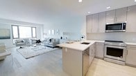 This single-floor space showcases a thoughtfully designed open-concept layout that seamlessly integrates the kitchen and living area into one cohesive environment. The kitchen is outfitted with sleek, flat-front cabinetry in a warm gray tone, paired with bright white countertops that extend into a peninsula with an integrated sink. The peninsula serves as both a functional workspace and a natural divider between the kitchen and the living area, enhancing the sense of flow throughout the space. Stainless steel appliances, including an oven and microwave, complement the modern aesthetic. Recessed lighting throughout keeps the space well-illuminated while maintaining a clean ceiling line. The living area features light-toned hardwood flooring that runs continuously from the kitchen, creating a unified and airy feel. A large window allows natural light to filter in, brightening the soft, neutral color palette of whites and light grays that defines the overall ambiance. The living space comfortably accommodates a seating arrangement anchored by a plush area rug, contributing to a calm and inviting atmosphere throughout this well-appointed space.