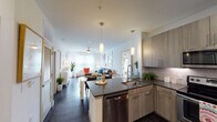 This single-floor residence is designed with a clean, contemporary aesthetic and a thoughtful open-concept layout. The kitchen serves as the heart of the home, showcasing wood-toned cabinetry, dark countertops, a white subway tile backsplash, and a full suite of stainless steel appliances including an electric range, over-the-range microwave, and French door refrigerator. A central island with a built-in sink and dishwasher provides additional prep space and connects seamlessly to the living area. Rich dark hardwood-style flooring runs throughout the main living spaces, grounding the bright, white walls and adding warmth to the interior. The living room is bathed in natural light from large windows dressed with sheer curtains and blinds, creating an inviting and airy atmosphere. Two bedrooms offer comfortable retreats, each featuring ceiling fans, plush carpeting, and generous windows that fill the rooms with natural light. The bathroom is well-proportioned, with large-format light tile flooring, wood-toned cabinetry, a white countertop, and direct access to a walk-in closet. Throughout the space, modern pendant lighting and a neutral color palette contribute to a cohesive, contemporary ambiance suited for everyday living.
