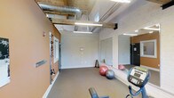 This single-level fitness space combines industrial and functional design elements to create a practical workout environment. The room features exposed ceiling ductwork and wooden beams overhead, giving it a raw, industrial aesthetic, while fluorescent strip lighting ensures the space is well-lit throughout. The walls are finished in a two-tone scheme, with a warm terracotta accent wall contrasting against neutral gray, lending visual interest to the utilitarian setting. Carpeted flooring provides a comfortable surface underfoot. Along the terracotta accent wall, wall-mounted resistance bands and suspension trainers offer versatile strength training options. A large mirror panel along the adjacent wall enhances the sense of space and allows users to monitor their form during exercise. Exercise balls of varying sizes, a dumbbell rack, and cardio equipment—including what appears to be a treadmill—round out the available workout tools. A window near the ceiling introduces a degree of natural light. A separate door leads to an adjoining room, suggesting additional facilities may be accessible nearby. Overall, the space is straightforward and efficiently equipped for a variety of fitness routines.
