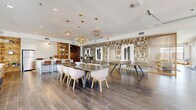 Located at 1226 Bexton St in Charlotte, NC, this thoughtfully designed single-floor clubhouse offers a dynamic mix of social and functional spaces. The open layout is anchored by large, black-framed floor-to-ceiling windows that flood the interior with natural light and provide views of the surrounding community and pool area.

The main gathering area showcases an eye-catching mosaic feature wall adorned with bold abstract artwork, complemented by a blend of sculptural pendant lighting — including woven and wire fixtures — that add warmth and visual interest. Dark wood-plank tile flooring runs throughout, grounding the contemporary aesthetic.

A spacious communal dining area features a substantial wooden table surrounded by modern upholstered chairs, ideal for group gatherings. Adjacent to it, a sleek kitchen island with white countertops and leather barstools serves as both a prep and social hub, flanked by built-in wood shelving with herringbone tile accents.

The lounge zones offer comfortable seating arrangements, including a tufted caramel leather sectional and neutral sofas set on layered rugs, creating an inviting atmosphere for relaxation. A bistro-style nook with counter stools sits beside a large window, perfect for casual conversation or individual use.

The space also includes a game table area and open co-working zones, making it a versatile hub for both leisure and productivity.