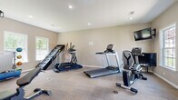 This dedicated fitness center offers a well-organized and fully equipped space designed to support a wide range of workout routines. The room features neutral-toned walls and durable carpet tile flooring, creating a clean and functional environment. Recessed lighting throughout the ceiling provides consistent, even illumination, while multiple windows along the walls invite natural light and offer views of surrounding greenery.

The cardio section is well-stocked with a Landice treadmill, an additional treadmill with an integrated console, an elliptical trainer, a stair climber, and a recumbent-style cycle. For strength training, the space includes a multi-functional cable machine with an adjustable bench, a rack of dumbbells in varying weights, and a selection of medicine balls and exercise balls for functional fitness. A wall-mounted flat-screen television is also present, providing entertainment during workouts.

The layout is thoughtfully arranged to allow ample space between equipment, ensuring comfortable movement throughout the room. A door provides access to an adjacent area, and informational fitness charts are displayed on the walls for user reference. This fitness center delivers a comprehensive and practical workout environment suited to a variety of fitness levels and training goals.