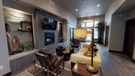 Located at 13101 Shetland Lane in Spokane, WA, this single-floor clubhouse facility delivers a refined and contemporary atmosphere throughout. The welcoming lounge area serves as the centerpiece of the space, anchored by a striking floor-to-ceiling stacked-stone fireplace wall with a wall-mounted TV, complemented by plush seating, designer pendant lighting, and large windows that invite natural light and outdoor views. Adjacent to the lounge, the kitchen is appointed with rich dark cabinetry, stainless steel appliances including a gas range and French door refrigerator, a mosaic tile backsplash, hexagonal tile flooring, and an expansive dark granite breakfast bar illuminated by a row of cube pendant lights — ideal for entertaining or casual gatherings. A dedicated office space features bold geometric-patterned wallcovering, wraparound windows offering neighborhood views, recessed lighting, and a substantial wood desk with ample seating. The corridor area connects to a fitness room and a restroom, finished with slate-style tile flooring and tall decorative foliage. A separate media room rounds out the amenities, outfitted with a large wall-mounted screen, leather sectional seating, a bold abstract artwork, and recessed ceiling lighting that creates a cozy, immersive environment. Throughout, the space maintains a cohesive, upscale design aesthetic with warm neutral tones and dark wood accents.