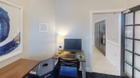 This single-floor business center is designed to provide a productive and visually engaging workspace. The room features neutral walls painted in soft white and light grey tones, creating a clean and professional atmosphere. The flooring transitions between patterned dark carpet and light tile near the entryway, adding visual interest throughout the space.

Multiple wooden workstations are arranged throughout the room, each paired with cushioned cross-back chairs upholstered in blue fabric. The workstations are equipped with desktop computers, keyboards, and task lighting via modern table lamps, ensuring a fully functional environment for productivity. A multifunction printer is also available, positioned conveniently along one of the workstation surfaces.

Natural light enters the room through a window fitted with white horizontal blinds, complementing the ambient lighting from the table lamps. A black multi-pane glass door provides access to the space, adding an architectural accent while allowing views to the exterior.

The walls are adorned with striking large-scale abstract artwork featuring bold blue brushstroke compositions in white frames, lending a contemporary and creative flair to the otherwise utilitarian setting. Two additional framed blue abstract prints are displayed vertically, reinforcing the cohesive color palette and polished aesthetic of the space.