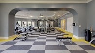 This fitness center is thoughtfully designed to support a full range of workout routines across two distinct zones connected by an elegant arched passageway. The main strength and cardio area features a bold checkerboard carpet in grey and white, accented by a vivid yellow baseboard that adds a dynamic energy to the space. The curved treadmill alcove, lined with a wraparound bank of windows, fills the room with natural light and offers views of the surrounding outdoor greenery. This area also houses elliptical machines, weight benches, a full rack of dumbbells, a cable machine, and a multi-functional power rack, all beneath recessed lighting and crown molding. A wall-mounted flat-screen TV and large mirrors enhance the experience. Through the archway, the cycling studio offers a bright, welcoming atmosphere with light hardwood-style flooring, ceiling fans, and multiple windows overlooking lush landscaping. The studio is outfitted with a fleet of stationary spin bikes, balance trainers, medicine balls, and a wall-mounted display screen for guided workouts. Tasteful framed artwork, including a bicycle-themed piece, adds a stylish touch. Together, the two zones deliver a comprehensive, well-appointed fitness experience in a clean and polished environment.