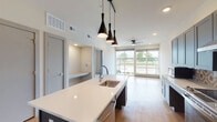 This single-floor residence showcases a thoughtfully designed open-concept layout that seamlessly connects the kitchen, dining, and living areas. The kitchen serves as a focal point, featuring a large central island with a white quartz countertop, an undermount sink, and a trio of sleek black pendant lights suspended from a track rail above. Cabinetry in a muted blue-gray tone lines the walls, complemented by a decorative patterned tile backsplash, stainless steel appliances including a range, refrigerator, and built-in microwave, and ample upper and lower storage. Light wood-style flooring extends throughout the main living areas, contributing to a clean, contemporary aesthetic.

The living area benefits from generous natural light provided by a wide floor-to-ceiling window wall that opens to a balcony, offering outdoor views. A ceiling fan is centrally positioned in this space. The bedroom features neutral carpet flooring, a ceiling fan with recessed lighting, and a side window. The bathroom includes a floating navy vanity with a white countertop, wood-look flooring, tiled tub surround, and a well-lit vanity mirror. The overall ambiance is modern, bright, and cohesive throughout.