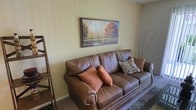 This single-floor space features a living area centered around a classic brown leather sofa accented with warm terracotta and patterned throw pillows, contributing to a welcoming and comfortable ambiance. The walls are finished in a soft, muted yellow-green tone that complements the earthy color palette throughout the room. A framed landscape painting depicting autumn foliage hangs above the sofa, serving as a focal point that reinforces the warm, nature-inspired aesthetic. A decorative arrangement of bare branches in a tall vase adds an organic, textural element to the corner of the room. To the left, a wooden ladder-style shelf provides both storage and visual interest. The flooring features a patterned area rug that grounds the seating area and adds additional depth to the design. The overall ambiance is casual yet thoughtfully arranged, with cohesive warm tones and carefully selected décor creating a space that feels both livable and aesthetically considered.
