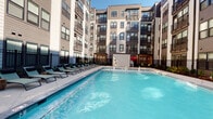 This single-floor unit is situated within a modern, multi-story residential building characterized by a contemporary exterior combining neutral siding, dark trim accents, and private balconies visible across multiple levels. The building's design centers around a well-appointed courtyard pool area, which serves as a prominent communal amenity. The outdoor pool is generously sized, featuring clear blue water, built-in water features, and ADA-accessible pool lift equipment. The pool deck is lined with lounge chairs along one side, and pergola-style shade structures add architectural interest to the surrounding hardscape. Landscaped planting beds with ornamental grasses border the fenced pool enclosure, which is framed on multiple sides by the building's façade, creating an enclosed, resort-like atmosphere bathed in natural sunlight.

Inside, the unit includes a bathroom finished in warm cream-toned walls with wood-accented fixtures throughout, including an ornate towel ring, an antique brass towel bar, and matching brass switch and outlet plates that lend a traditional character to the space. The bathroom features a vanity with a white sink, wood cabinetry, a framed mirror, and a full toilet. The overall interior palette is understated and cohesive.