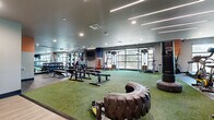 This expansive fitness center is thoughtfully designed to accommodate a wide range of training styles and fitness goals. The facility is organized into distinct zones, each with its own flooring to reinforce its purpose. A generous artificial turf area runs through the heart of the space, serving as the functional training zone and housing heavy-duty equipment such as a large tire for flipping drills, a sled, and freestanding punching bags — ideal for high-intensity functional workouts. Adjacent to this zone, rubberized flooring supports a well-stocked strength training area outfitted with a full dumbbell rack, multiple weight benches, squat racks, and cable machines. A dedicated cardio section lines the perimeter and features rows of treadmills, stationary bikes, ellipticals, and stair climbers, many positioned in front of large floor-to-ceiling windows that invite natural light and offer outdoor views. Wall-length mirrors amplify the sense of space throughout. The facility is illuminated by a combination of sleek linear LED strip lighting and recessed can lights set against a modern grey ceiling, creating a bright, energizing atmosphere. Exposed concrete accents and bold orange wall details add an industrial yet dynamic character to the overall aesthetic.