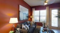 This single-floor space makes a strong visual impression through its bold design choices and thoughtful layout. The living area is defined by rich, deep red accent walls that create a warm and inviting ambiance, complemented by a large-scale abstract canvas artwork that serves as a striking focal point. A ceiling fan with an integrated light fixture adds both functionality and a casual elegance overhead. Natural light flows generously into the room through a prominently positioned window dressed with sheer white curtains and horizontal blinds, offering a glimpse of lush greenery beyond — a refreshing connection to the outdoors. The living space is furnished with a plush, charcoal-toned sofa adorned with an eclectic mix of patterned throw pillows, adding depth and texture to the room. A rich, dark shag area rug anchors the seating area, enhancing the sense of comfort and warmth. Dual floor lamps on either side of the sofa provide layered lighting, creating a balanced and welcoming glow throughout the space. The overall ambiance is one of curated, contemporary style — bold in color, rich in texture, and thoughtfully designed to feel both lived-in and visually dynamic.