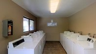 This shared laundry facility is a practical, single-floor space designed to accommodate multiple users efficiently. The room is laid out with two parallel rows of top-loading washing machines and dryers lining both walls, providing ample capacity for residents or guests. The machines are arranged in a clean, organized fashion, maximizing the use of the available floor space.

The interior features durable stone-tile flooring in warm earth tones, offering a hard-wearing surface well-suited to the demands of a high-traffic utility space. The walls are finished in a neutral taupe tone, giving the room a clean and understated appearance. Overhead, a large flush-mount fluorescent light fixture provides bright, even illumination throughout the space.

A large window on one wall allows natural light to filter in, enhancing the overall brightness of the room and providing a view of the outdoor surroundings. An informational notice board is mounted on the wall, likely displaying usage guidelines or instructions for users of the facility.

Overall, the laundry room presents a functional and well-maintained environment, thoughtfully arranged to serve the needs of a shared-use setting. Its straightforward layout, adequate lighting, and generous number of machines make it a convenient and efficient amenity.