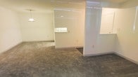 This single-floor space presents a clean, open layout characterized by neutral cream-colored walls and consistent grey carpeting throughout. The interior is divided into distinct zones using partial partition walls, which create a sense of separation while maintaining an open, connected feel across the space. The partitions feature a high-gloss white finish, adding a modern contrast to the warmer wall tones and providing visual interest without fully enclosing any area.

Recessed and overhead fluorescent lighting illuminates the space evenly, reflecting off the glossy partition surfaces and contributing to a bright, functional atmosphere. A pass-through window is visible in the background, suggesting a dedicated counter or service area that connects adjacent zones. The flooring transitions subtly near the partition base, hinting at a defined entryway or transition point between areas.

The layout appears well-suited for a professional or commercial use, with its straightforward design emphasizing practicality and ease of movement. The overall ambiance is understated and functional, with a neutral palette that allows for flexible customization to suit a variety of purposes.
