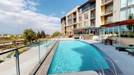 This outdoor amenity level showcases a thoughtfully designed resort-style environment set against the backdrop of a contemporary multi-story residential building. The centerpiece is a generously sized lap-style swimming pool with striking blue water, framed by sleek glass-panel railings that preserve the open, airy feel of the space. Adjacent to the pool sits a bubbling hot tub, elevated on a wide concrete surround and accompanied by cushioned lounge chairs shaded by bold orange cantilever umbrellas — ideal for relaxation in the sun.

A fully equipped outdoor kitchen anchors one end of the deck, featuring dual stainless steel grills, granite countertops, a utility sink, and ample cabinetry — all sheltered beneath a modern pergola structure. Nearby, alfresco dining and seating areas offer round wood-topped tables and wicker lounge sets, surrounded by curated ornamental plantings that add color and texture to the hardscape.

The elevated terrace includes a gated walkway that opens to panoramic views of a nearby greenway corridor and distant mountain peaks, lending the space a strong connection to the natural landscape. The overall design balances urban sophistication with outdoor comfort, creating a cohesive and functional amenity space suited for both lively gatherings and quiet leisure.