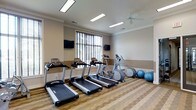 This fitness center is designed to provide a functional and comfortable workout environment. The space features a striped hardwood-style floor that adds a polished, clean aesthetic to the room. Crown molding lines the upper walls, lending a refined architectural detail that elevates the overall appearance beyond a standard gym setting.

The cardio area is well-stocked, featuring multiple treadmills and an elliptical trainer positioned toward the center of the room. An upright stationary bike and additional cardio equipment round out the selection. Exercise balls are neatly arranged along one wall, providing options for stretching and core workouts.

Two flat-screen televisions are wall-mounted, allowing users to stay entertained during their workouts. A large window fitted with horizontal blinds brings in natural light, brightening the space considerably. A ceiling fan overhead helps maintain airflow and comfort during exercise sessions.

Recessed fluorescent lighting fixtures are evenly distributed across the ceiling, ensuring consistent illumination throughout the room. The neutral beige and cream wall tones create a calm, welcoming atmosphere, making this fitness center a practical and inviting space for daily exercise.