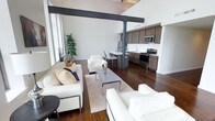 This two-floor space makes a striking first impression with its soaring ceilings and bold exposed steel beams that span the open-concept main living area, lending an industrial-chic character to the overall design. Rich, dark hardwood flooring runs throughout, grounding the bright, airy atmosphere created by the generous vertical space above.

The living area is expansive and thoughtfully arranged, with ample room for multiple seating configurations. Clean-lined white upholstered sofas and chairs surround a sleek chrome-framed coffee table set atop a light area rug, creating a comfortable and contemporary gathering space. Tall windows along the perimeter allow natural light to filter in, while a potted indoor tree adds a touch of organic warmth.

The kitchen flows seamlessly from the living area and is outfitted with medium-toned wood cabinetry, stainless steel appliances including a range and microwave, and a clean white countertop workspace. A raised breakfast bar with bar seating serves as a natural dividing element between the kitchen and the living space, making it ideal for casual dining or entertaining.

The second floor adds additional living or sleeping space, complementing the generous layout of the main level. The overall ambiance is modern, open, and sophisticated, with industrial details balancing clean contemporary finishes.