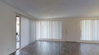 This single-floor space offers a clean and functional layout designed for comfortable everyday living. The main living area is the centerpiece of the space, showcasing smooth wood-style flooring that runs throughout, lending warmth and a modern aesthetic to the interior. A large picture window dressed with vertical blinds dominates one wall, allowing an abundance of natural light to fill the room while providing privacy and light control as needed. The textured ceiling adds a subtle visual layer to the otherwise clean, neutral palette of white and soft gray tones that define the space. A secondary entry door is conveniently positioned adjacent to the main window, offering practical access. The open and unobstructed floor plan provides flexibility in furniture arrangement, making it adaptable to a variety of uses and personal styles. The overall ambiance is bright, airy, and inviting, with a minimalist character that serves as a versatile canvas for personalization. The combination of natural light, durable flooring, and a well-proportioned layout makes this a practical and welcoming space.