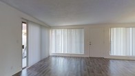 This single-floor space offers a clean and functional layout designed for comfortable everyday living. The main living area is the centerpiece of the space, showcasing smooth wood-style flooring that runs throughout, lending warmth and a modern aesthetic to the interior. A large picture window dressed with vertical blinds dominates one wall, allowing an abundance of natural light to fill the room while providing privacy and light control as needed. The textured ceiling adds a subtle visual layer to the otherwise clean, neutral palette of white and soft gray tones that define the space. A secondary entry door is conveniently positioned adjacent to the main window, offering practical access. The open and unobstructed floor plan provides flexibility in furniture arrangement, making it adaptable to a variety of uses and personal styles. The overall ambiance is bright, airy, and inviting, with a minimalist character that serves as a versatile canvas for personalization. The combination of natural light, durable flooring, and a well-proportioned layout makes this a practical and welcoming space.