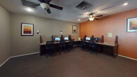 This single-story space is thoughtfully arranged to serve multiple functional purposes. The main computer lab is a generously sized room featuring rows of individual workstations, each equipped with desktop computers and partitioned dividers for privacy. Two ceiling fans with integrated lighting fixtures keep the room comfortable, while a bold orange accent wall adds a pop of color against the neutral taupe walls. Framed artwork provides decorative interest throughout. A smaller, semi-private workstation area is tucked into an adjacent alcove, also finished with the same warm orange accent wall, housing a single desktop setup with cabinetry below. A separate office area features a large window fitted with horizontal blinds, allowing natural light and outdoor views of a landscaped exterior. A ceiling fan, recessed lighting, and a corner desk with ergonomic seating make this a practical workspace. An additional lounge-style room includes a wall-mounted flat-screen TV, a framed landscape painting, and visible AV and data connections along the walls. Dark carpeting runs throughout the space, contributing to a consistent, professional aesthetic overall.
