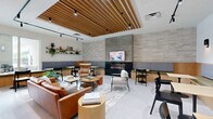 Located at 15955 NW 6th Avenue in Miami, FL, this thoughtfully designed communal lounge space offers a welcoming blend of modern aesthetics and functional comfort. The defining architectural feature is the striking slatted wood ceiling, which adds warmth and visual depth while framing the open floor plan below. Track lighting runs throughout, complementing the natural tones of the wood and the crisp white walls.

The space is organized into several distinct yet interconnected seating zones. A central lounge area features rich cognac leather sofas and accent chairs arranged around a rustic round coffee table atop a layered area rug, creating an inviting focal point. Adjacent to this is a dramatic floor-to-ceiling textured stone wall housing a linear electric fireplace below a wall-mounted television — ideal for gathering and relaxation.

Built-in banquette seating with wood-trimmed bases lines portions of the perimeter, paired with bistro-style tables and black chairs to accommodate casual dining or work. Decorative open shelving with curated greenery and accents adds personality to the walls. A recessed wet bar niche with dark countertops and wood cabinetry provides functional amenities. Large glass doors along the exterior flood the space with natural light and offer views of the outdoor surroundings, enhancing the airy, open ambiance throughout.