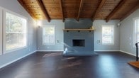 This two-story home showcases a thoughtful blend of rustic character and modern practicality. The main living area immediately impresses with soaring vaulted ceilings clad in warm wood planks and supported by exposed timber beams, creating an open, airy atmosphere. A bold, floor-to-ceiling brick fireplace with a wood mantel serves as the room's focal point, flanked by windows that invite natural light throughout. Dark hardwood flooring runs through the main level, complementing the earthy tones of the ceiling above. A striking open staircase with natural wood treads and black metal railings connects the two floors, adding an architectural element to the space. The kitchen features warm wood cabinetry, a slate-toned tile backsplash, white countertops, and black appliances, offering both style and functionality. A full bathroom on the main level includes a walk-in shower, while a second bathroom features a white vanity with ample counter space and hardwood-style flooring. Upstairs, carpeted bedrooms are bathed in natural light through multiple windows with outdoor views, and a loft-style area with an interior railing overlook showcases the exposed wood beam ceiling below, adding a sense of openness and connection between the two levels.