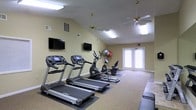 This single-floor fitness center offers a practical and inviting space for exercise. The room features vaulted ceilings with ceiling fans, contributing to a sense of openness and improved air circulation during workouts. Soft yellow walls create a light, welcoming atmosphere throughout the space.

The cardio area is equipped with multiple treadmills and stationary bikes, positioned to face two wall-mounted flat-screen televisions, allowing users to stay entertained during their workouts. Exercise balls and additional fitness equipment are visible along the walls, offering versatile training options.

Carpeted flooring runs throughout the space, providing a cushioned surface underfoot. A set of double glass doors at the far end of the room allows natural light to filter in while providing direct access to the outdoors. Recessed and overhead fluorescent lighting ensures consistent brightness throughout the room.

The layout is thoughtfully arranged to maximize usable floor space while maintaining clear pathways between equipment, creating a functional yet comfortable environment suitable for a variety of fitness routines.