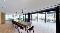 This upscale rooftop amenity space masterfully blends interior comfort with outdoor living through a series of expansive floor-to-ceiling glass walls and wide bi-fold doors that open fully to a spacious exterior terrace. The interior is anchored by a generously sized communal dining table crafted from reclaimed wood, surrounded by a mix of modern black and white chairs beneath elegant cone-shaped pendant lights. Herringbone-patterned brick flooring runs throughout the interior, adding warmth and texture to the contemporary design. A striking built-in shelving unit with Gothic arch detailing in natural light wood serves as a dramatic backdrop, displaying curated decorative objects and glassware. A cozy lounge area features a vibrant yellow sofa, armchairs, and a glass coffee table set atop a layered rug, framed by corner glass walls that invite abundant natural light and city views. The outdoor terrace is finished with large-format stone pavers and includes a pergola-covered seating area with lounge chairs, planters with greenery, and open sightlines to the surrounding urban skyline. Together, the indoor and outdoor areas create a versatile, design-forward environment ideal for gathering and entertaining.